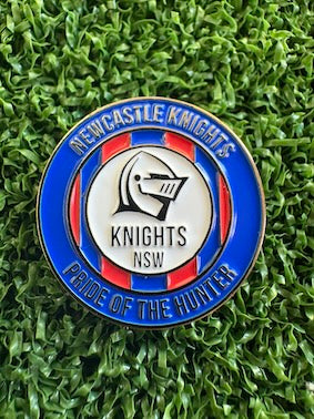 A circular Newcastle Knights NSW sports pin badge with a blue, red, and white enamel design, placed on a green grass background.